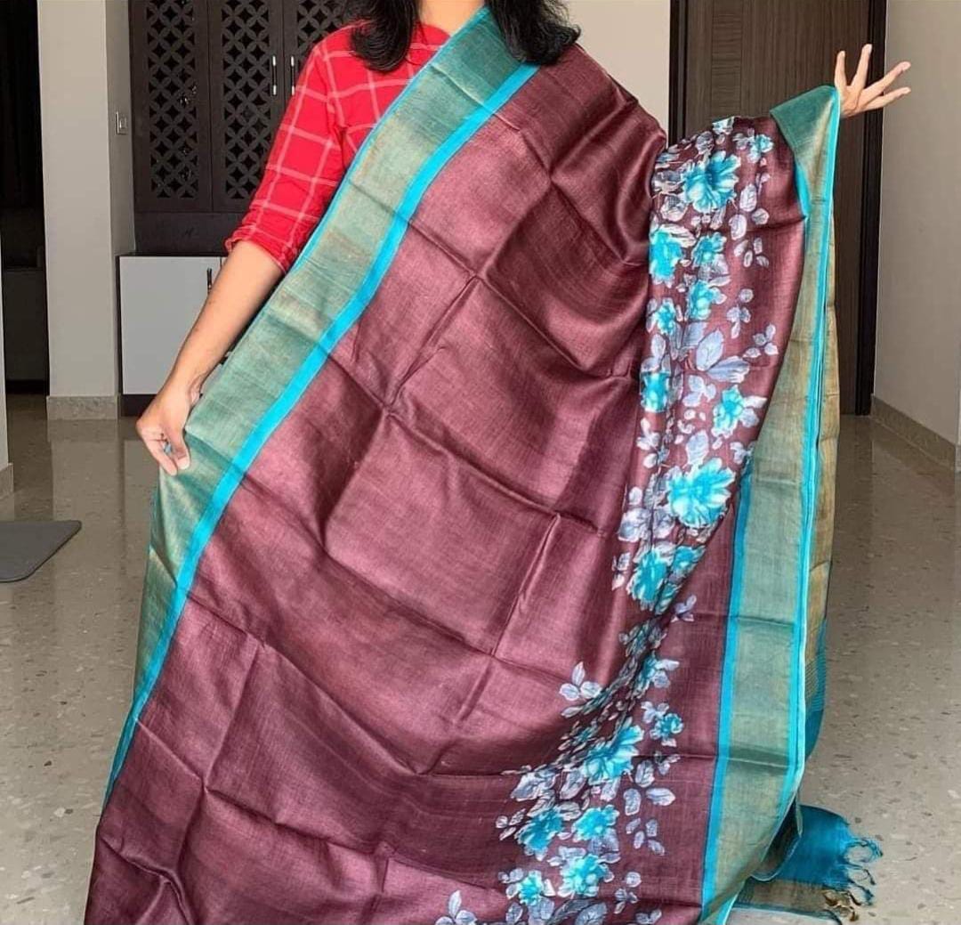 A woman holding a Handloom Bhagalpuri Tussar Saree with gold borders and a floral panel design in a color scheme that includes purple and blue.