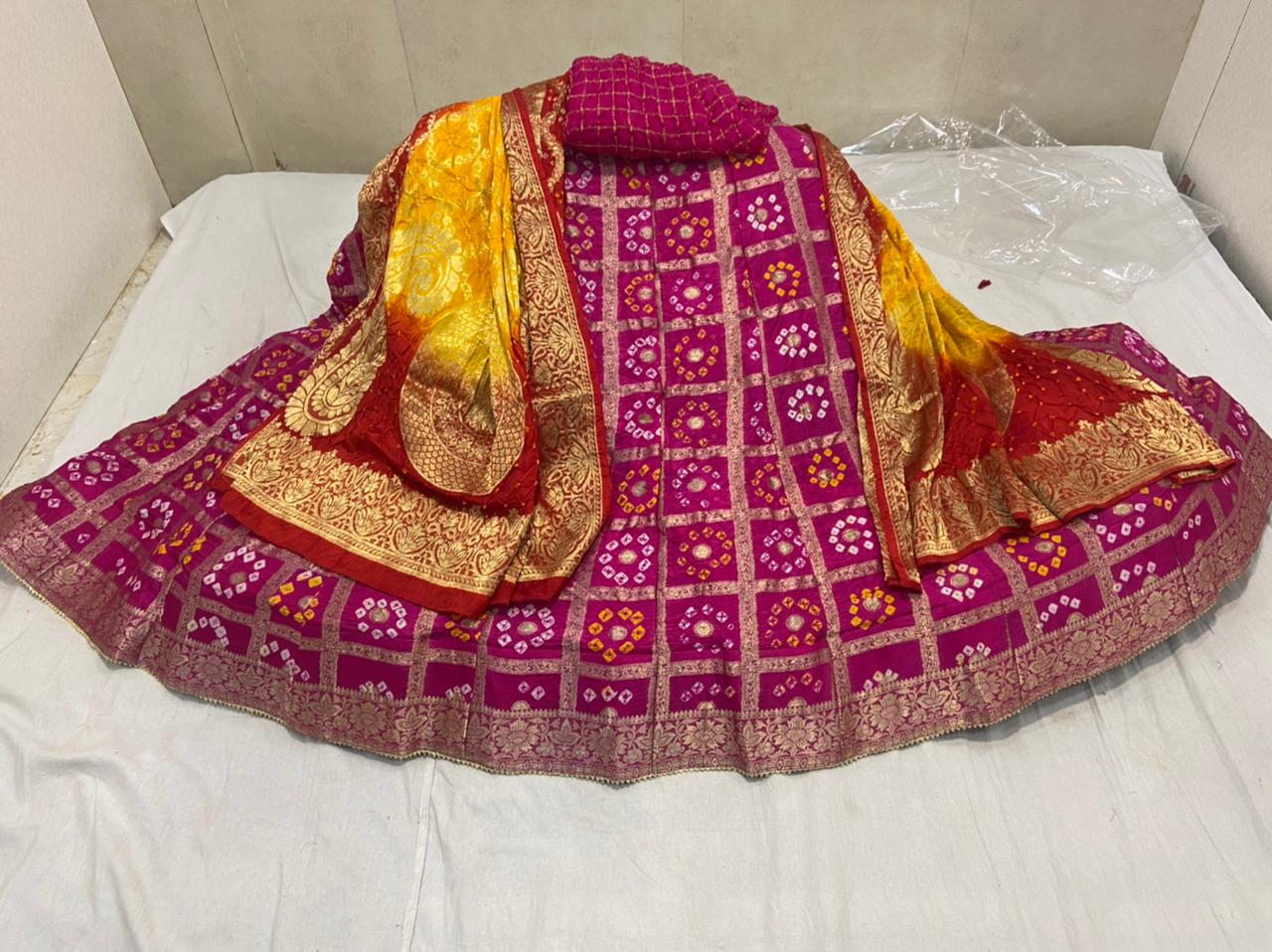 A Banarasi Gharchola silk lehenga set with a bandhani design, zari blouse, and dupatta, primarily in yellow and pink colors with floral and geometric patterns.