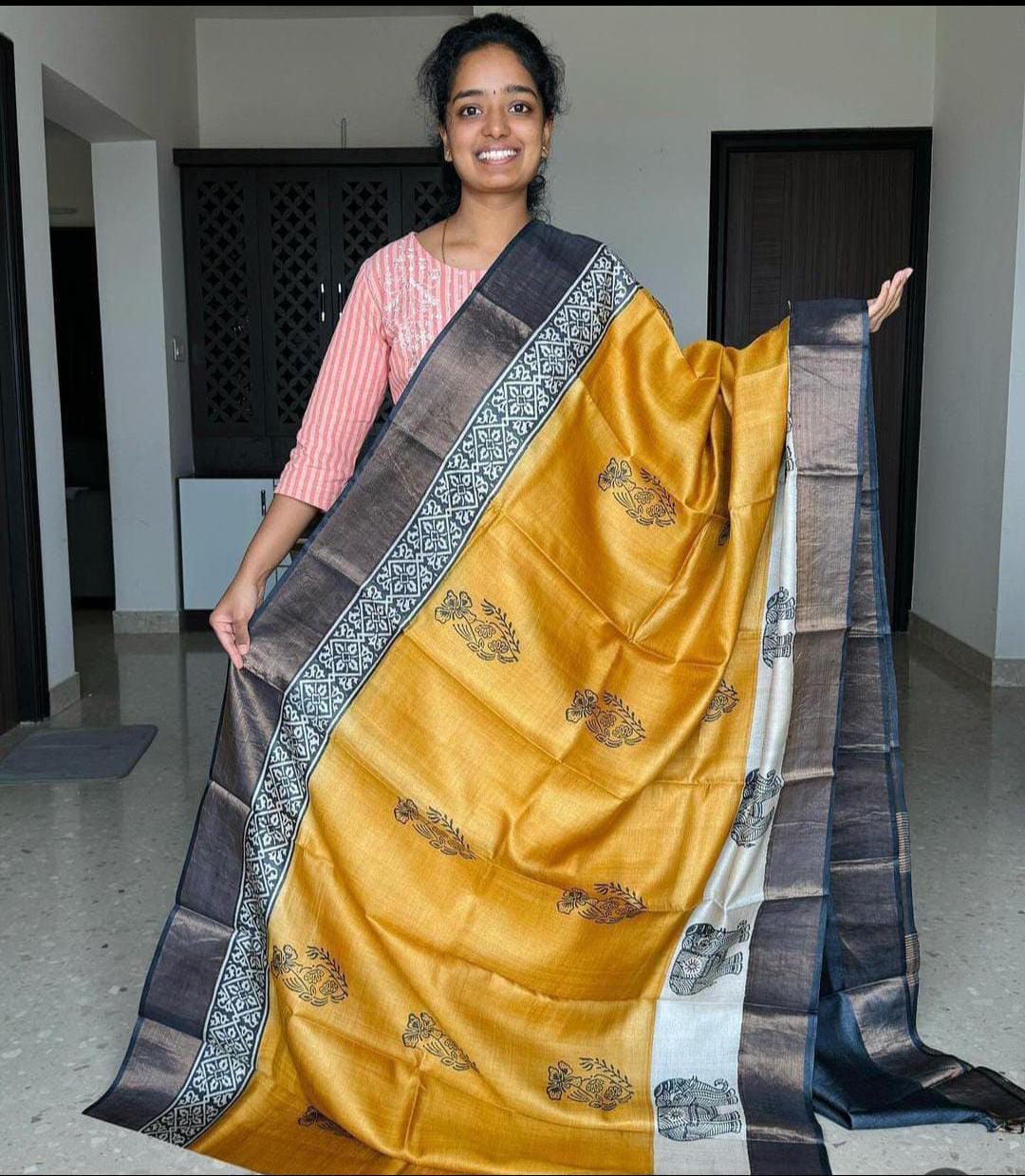 A woman holding a Handloom Bhagalpuri Tussar Saree with gold borders and intricate panel design in a home setting.