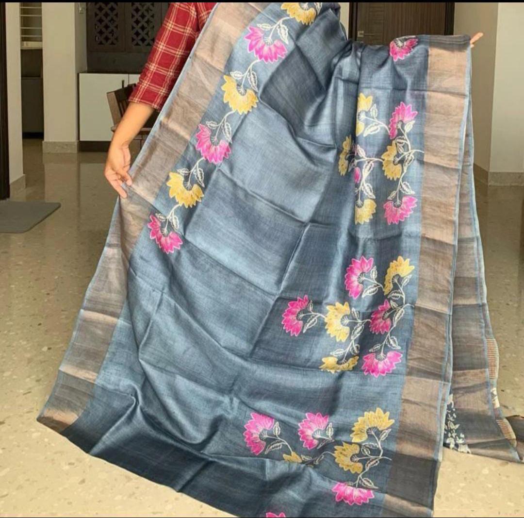A person holding a draped Handloom Bhagalpuri Tussar Saree with gold borders and a floral panel design in blue and beige colors.