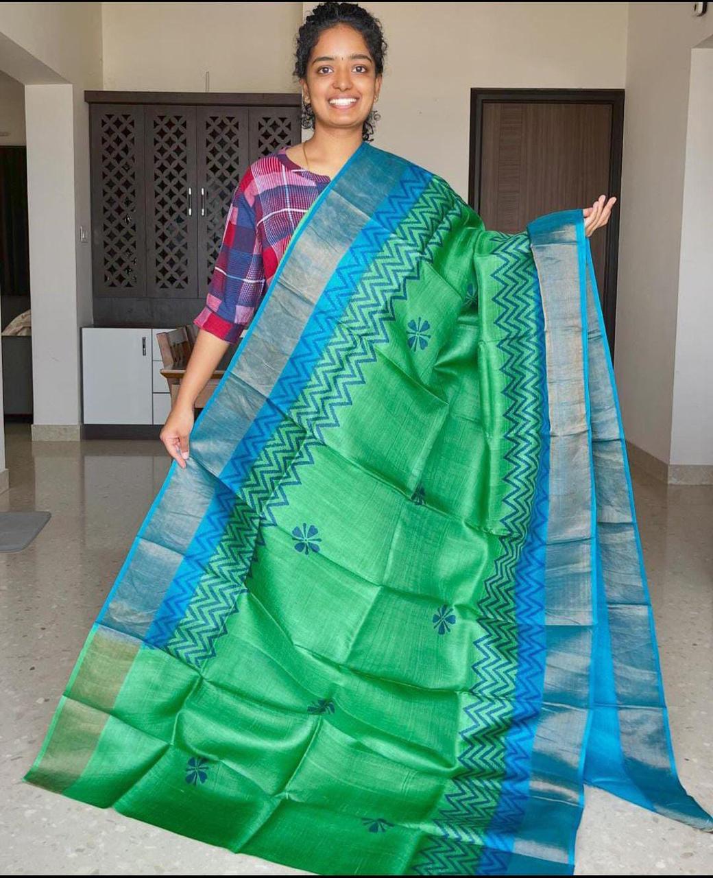 A woman smiling, holding a green and blue Handloom Bhagalpuri Tussar Saree with gold borders and a geometric panel design.