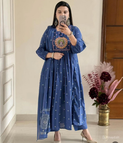 Woman wearing a blue embroidered dress taking a mirror selfie in a room with a vase of flowers.