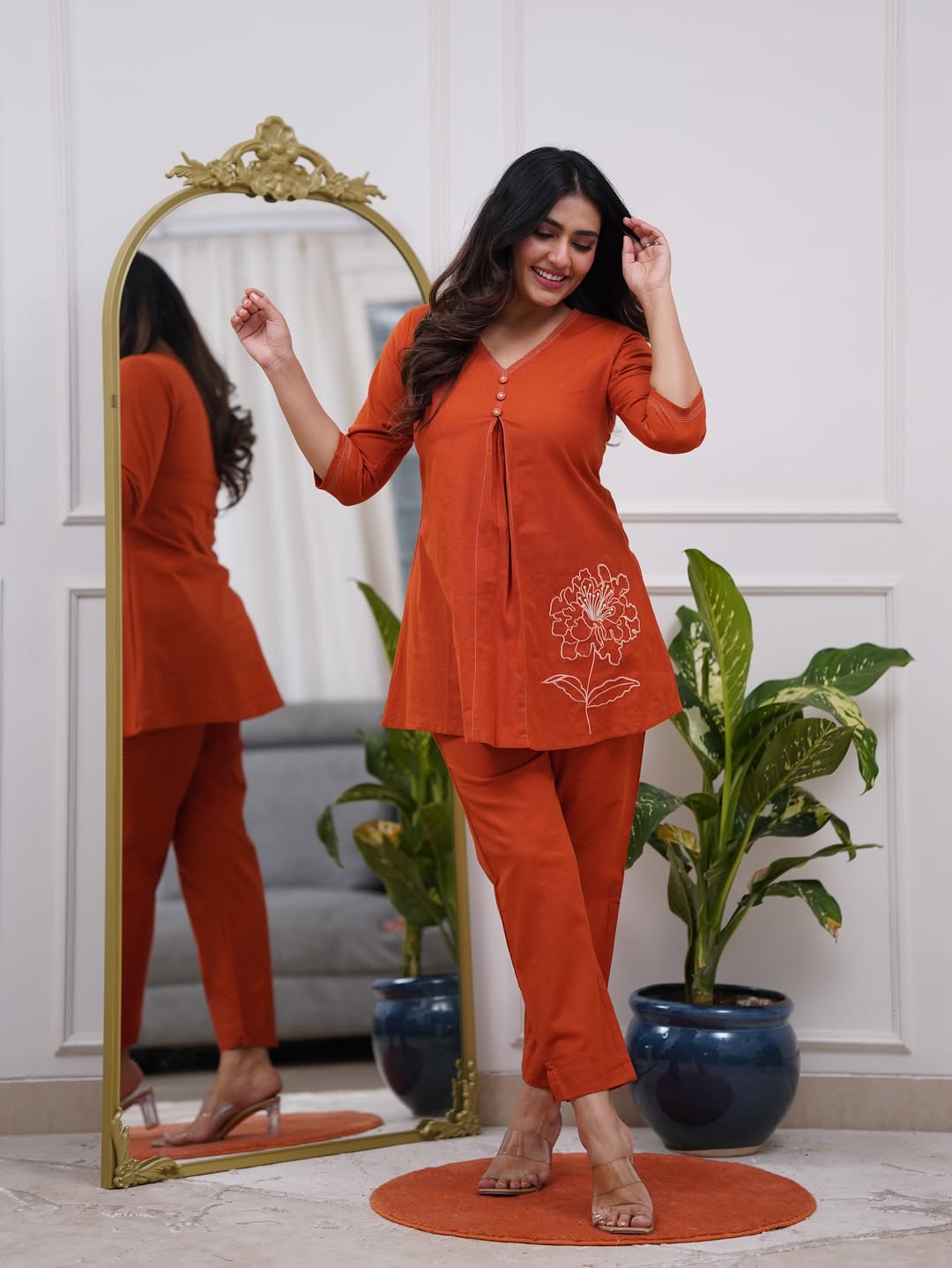 Woman in an orange outfit standing next to a full-length mirror in a room with plants.