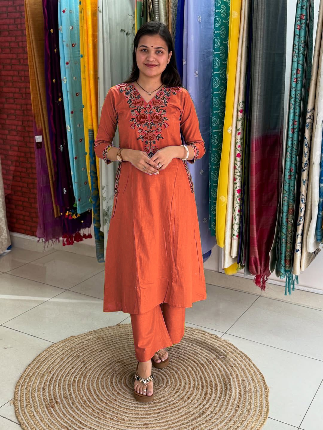 Woman in an orange traditional outfit standing in front of colorful fabric curtains.