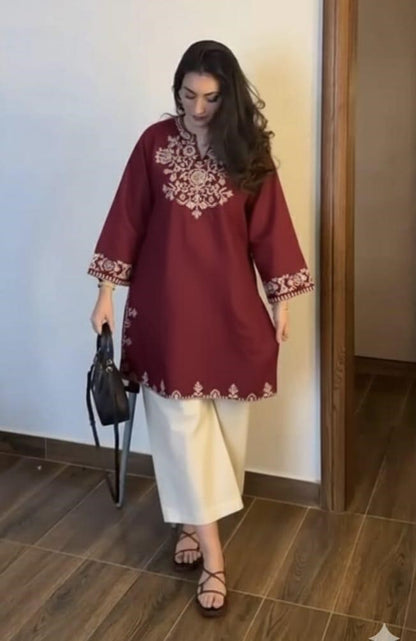 A woman wearing a maroon embroidered top and white pants is standing indoors.