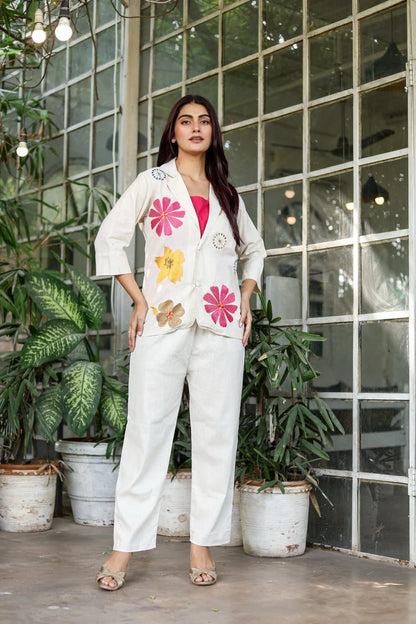 Woman in a white outfit with floral patterns standing in front of a glass wall with plants.