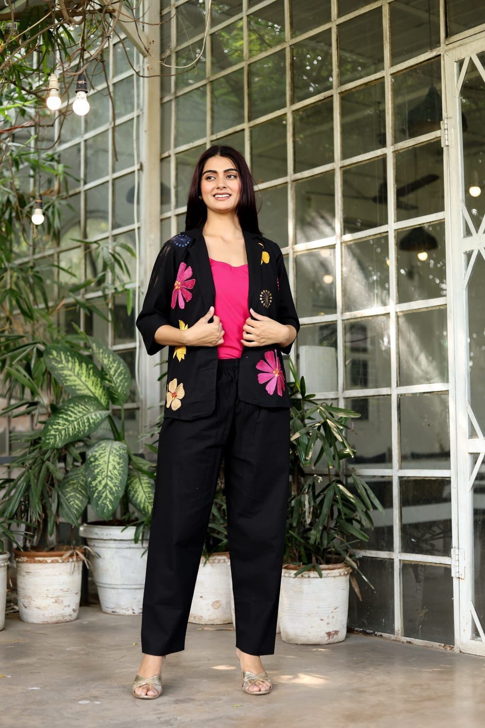 Woman in a black outfit with floral embellishments standing in front of a glass wall with plants.