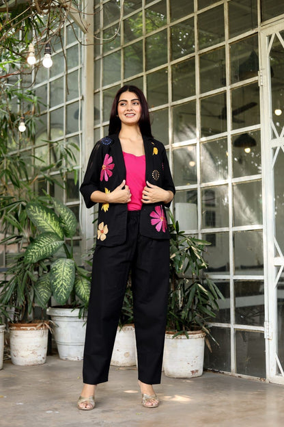 Woman in a black outfit with floral embellishments standing in front of a glass wall with plants.