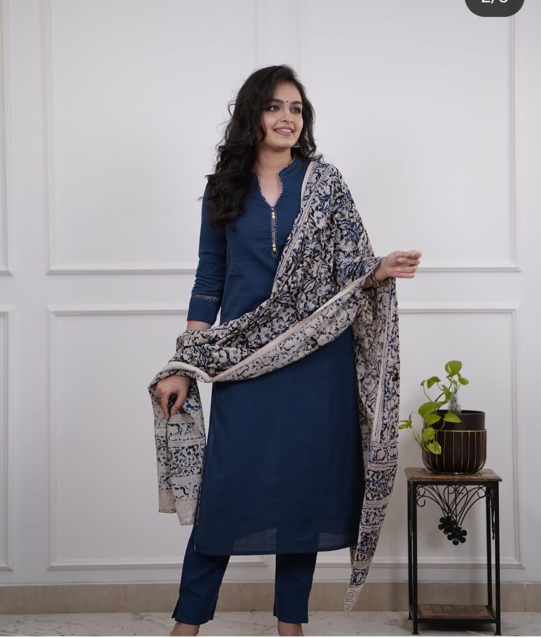 Woman wearing a blue outfit with a patterned shawl standing against a white paneled wall.