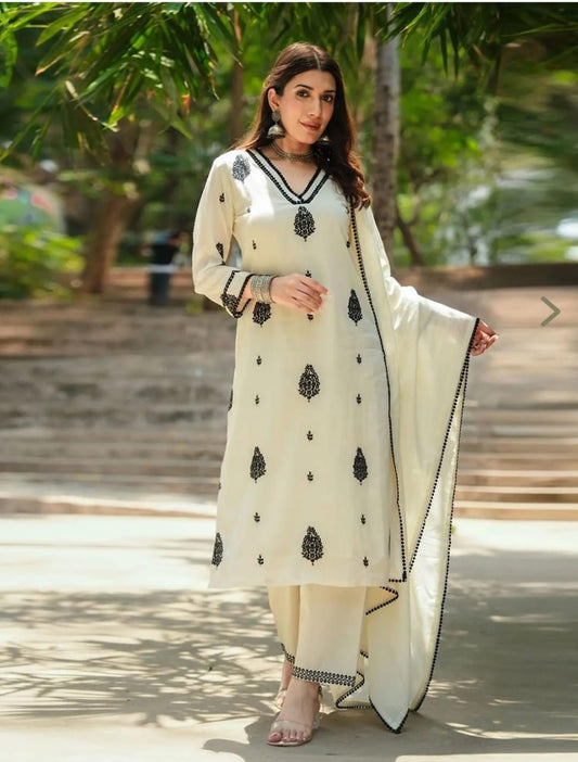 Woman in a cream-colored traditional outfit with black patterns standing outdoors.