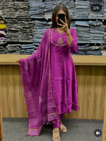 Person wearing a purple traditional outfit with gold embroidery in front of stacked fabric.