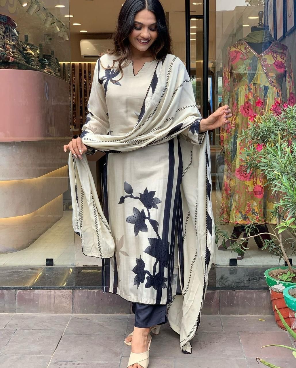 Woman in a floral outfit standing in front of a store with mannequins and plants.