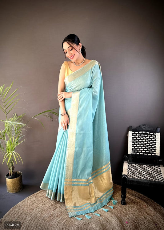 Woman wearing a light blue saree with a beige border, standing against a gray wall with a plant and chair in the background.