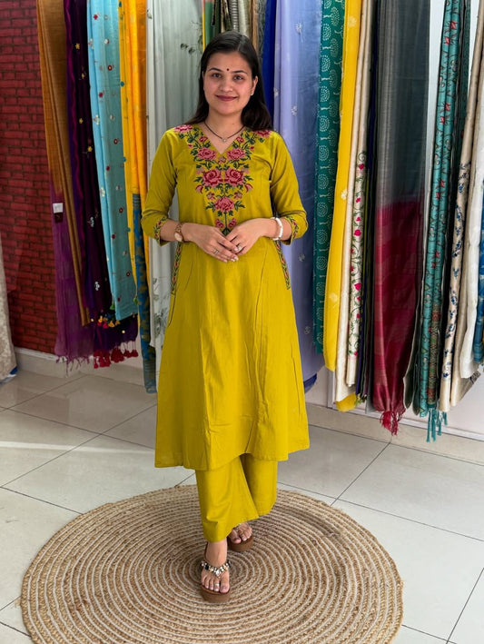 Woman in a yellow traditional outfit with floral embroidery standing in front of colorful fabric curtains.